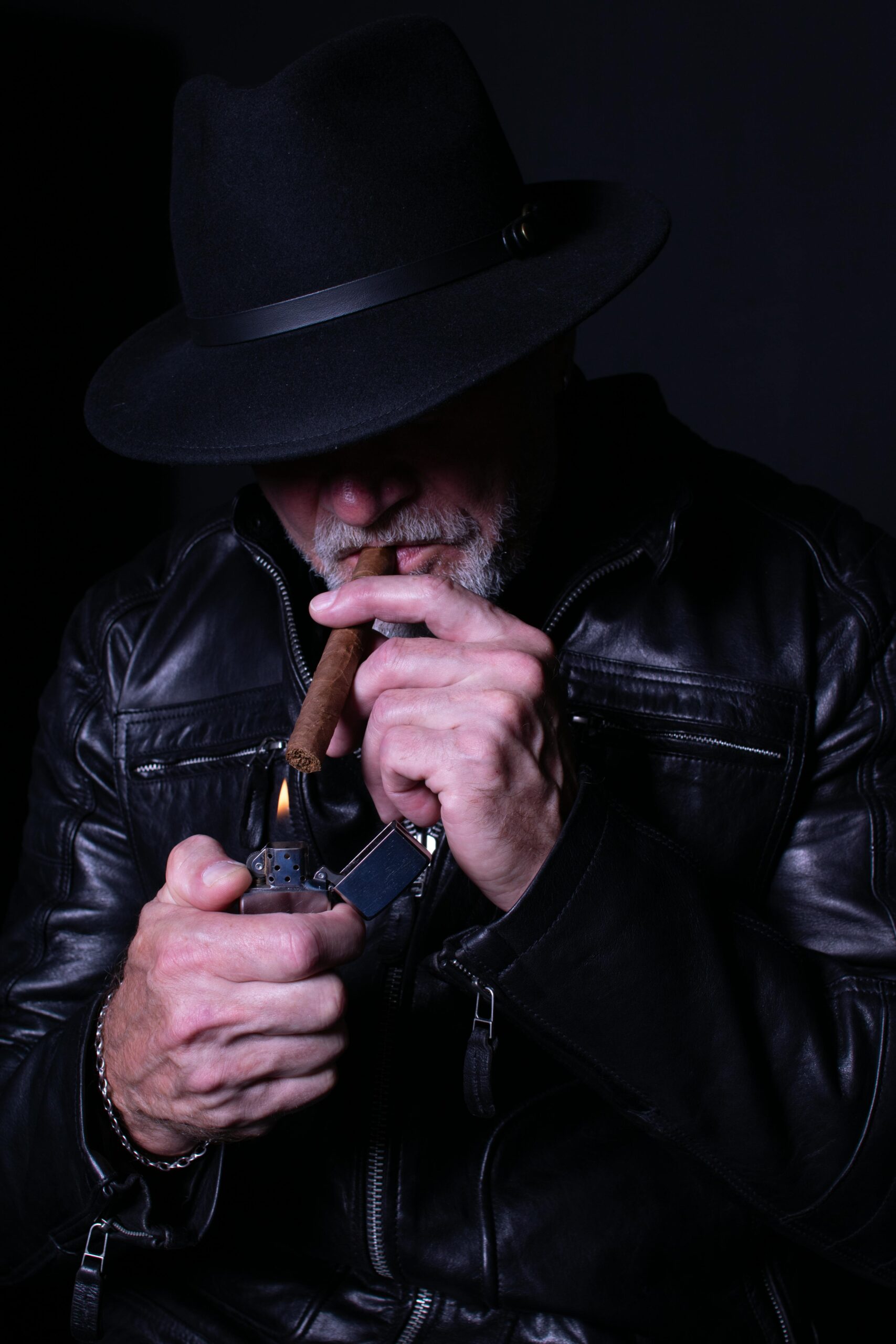 Senior man in leather jacket and hat lighting a cigar in a dark setting.