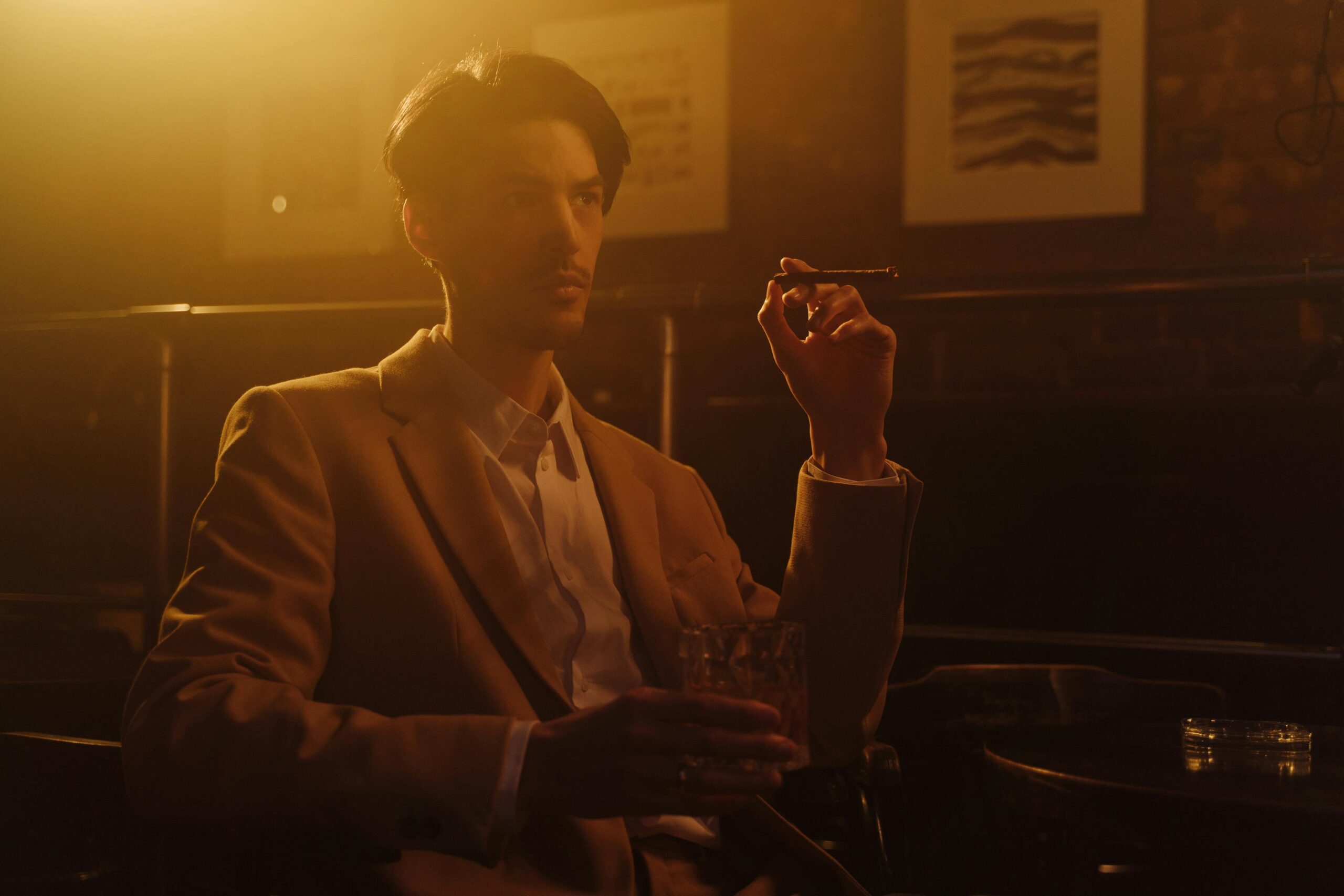 A man in a suit relaxes with a cigar and whiskey in an elegant setting.