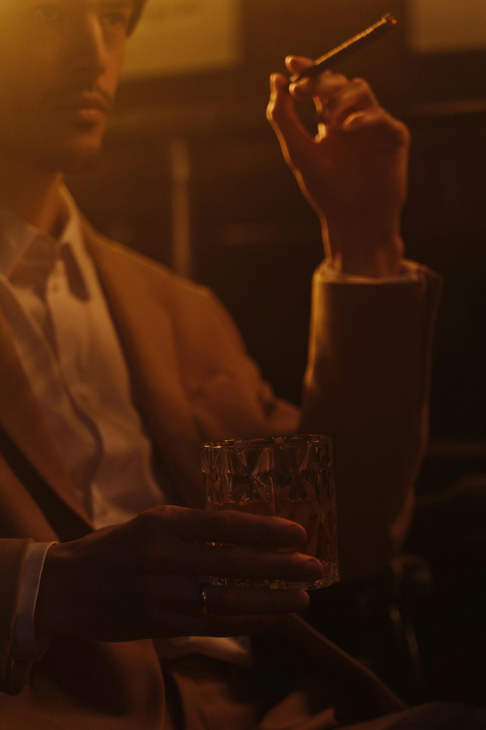 A contemplative man elegantly holding a whiskey glass and cigar in a warm, ambient setting.